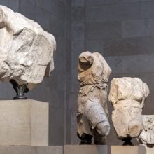 Three fragmented ancient marble sculptures, depicting torso figures draped in flowing garments, displayed on stone pedestals within a softly lit museum setting.