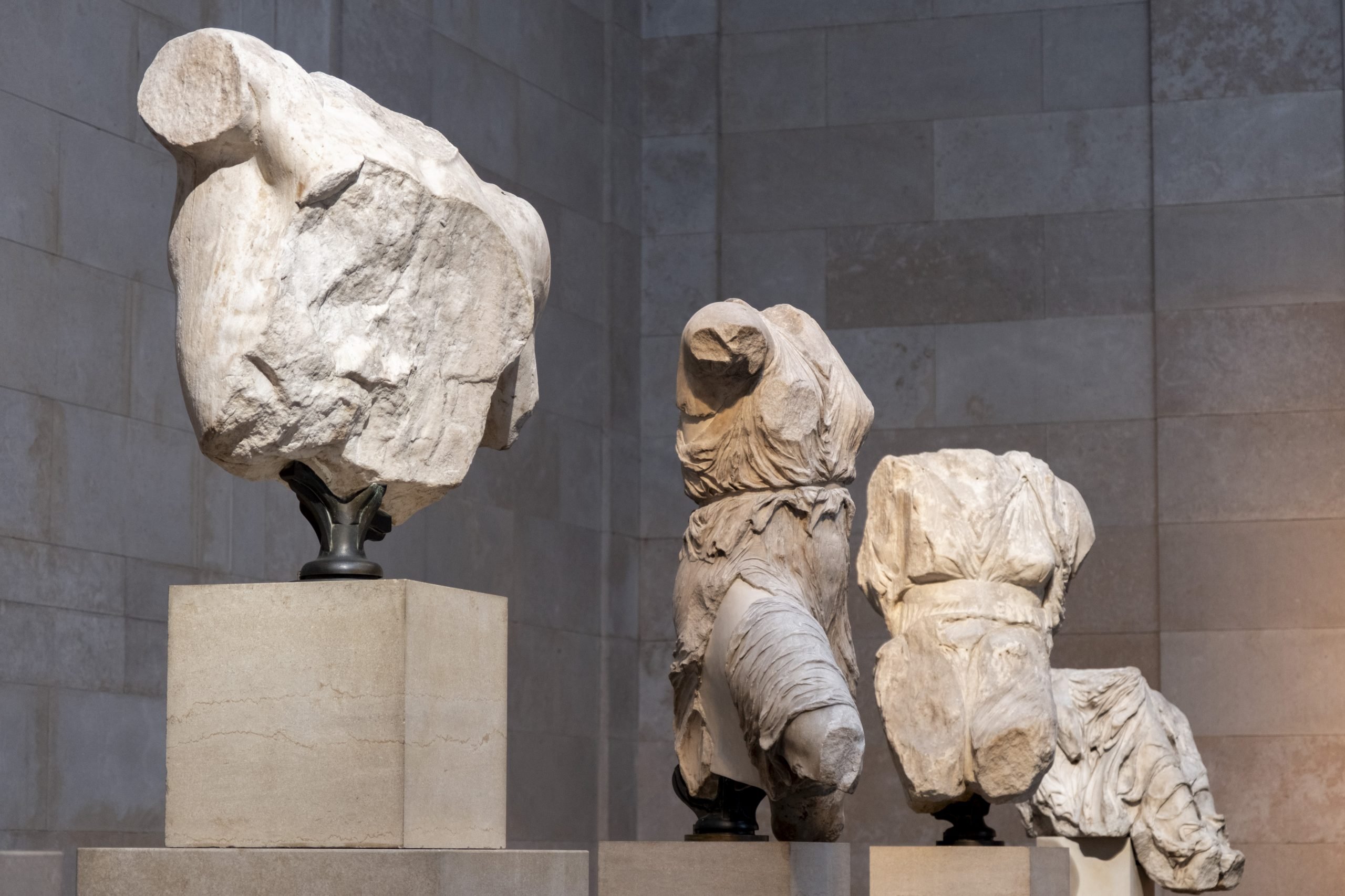 Three fragmented ancient marble sculptures, depicting torso figures draped in flowing garments, displayed on stone pedestals within a softly lit museum setting.