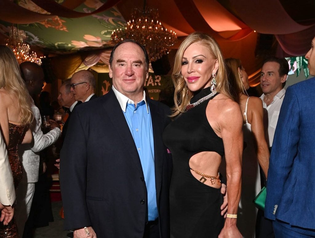 A well-dressed couple poses at an elegant evening event with chandeliers and vibrant draped ceilings, surrounded by other formally dressed guests.