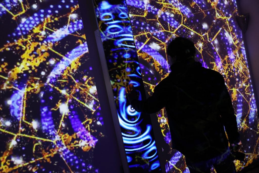 A person interacts with a vibrant digital art installation featuring swirling blue patterns and golden geometric designs illuminated on vertical panels.