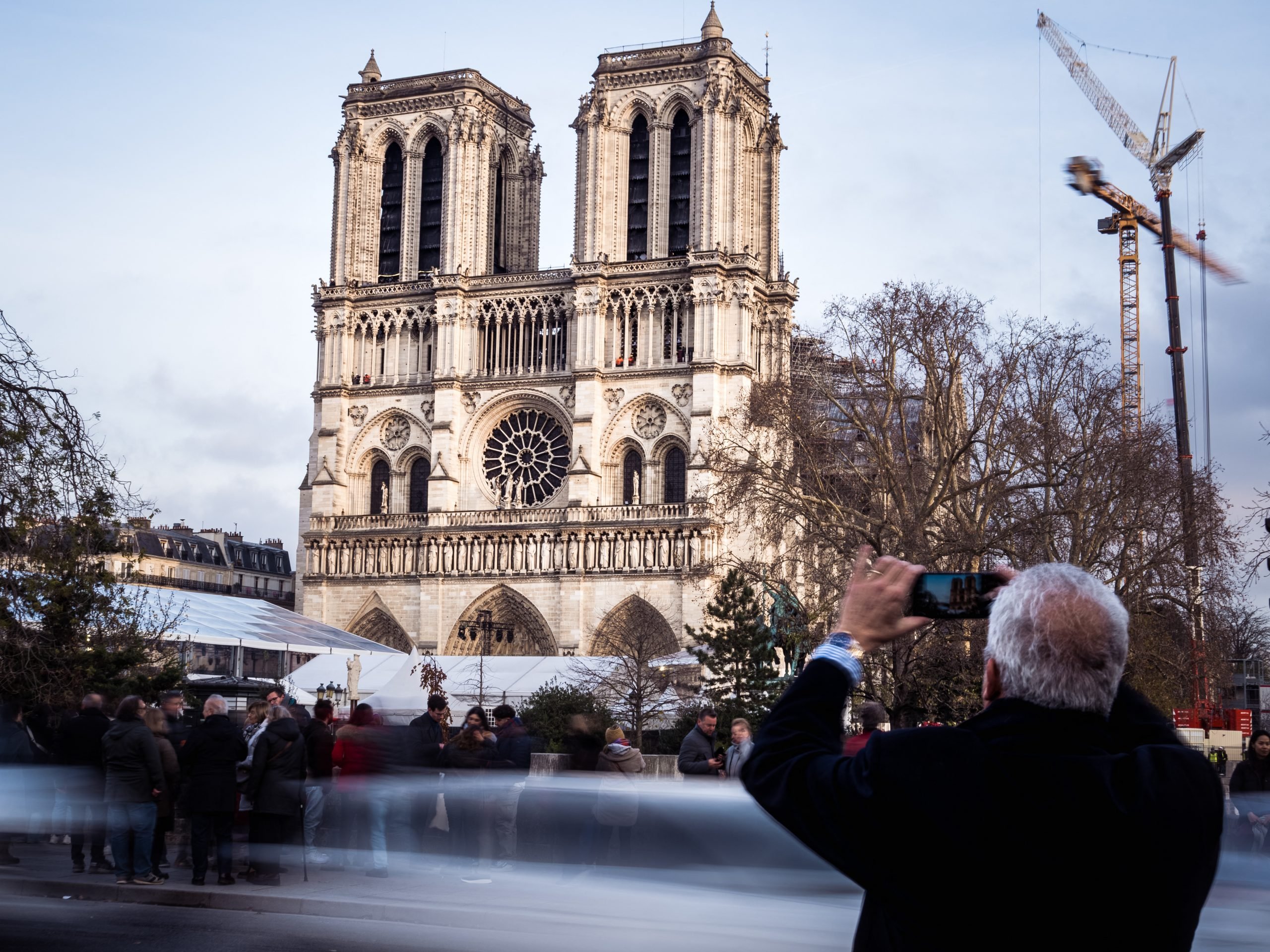 a man take a picture of notre dame cathedral as a crowd of people walks by in the foreground