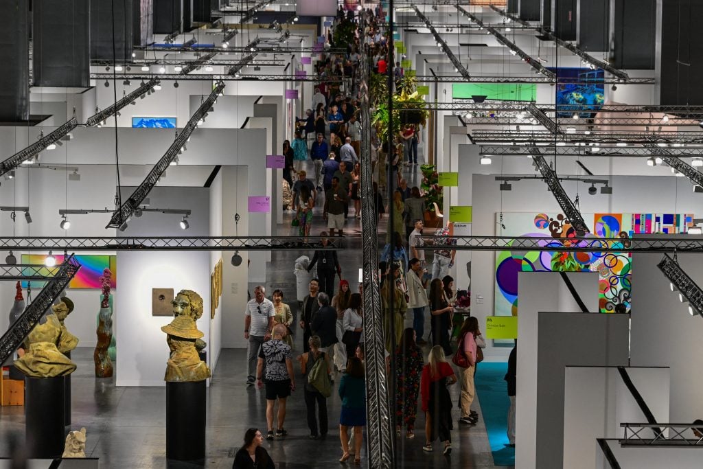 Many people walk around white-walled booths filled with art.