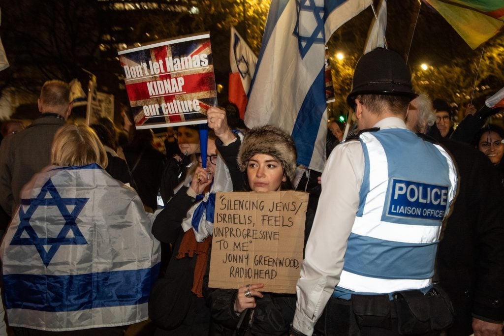 a police officer in a high viz vest stands in front of a group of protestors holding up signs and wearing Israeli flags