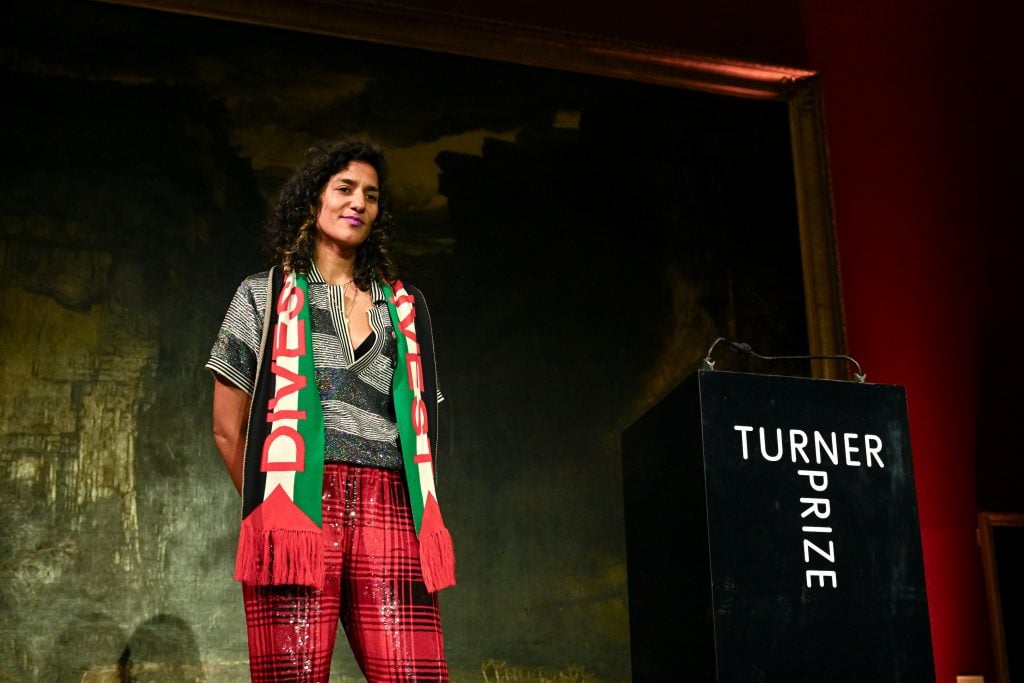 a woman in a red, green, and black scarf stands next to a podium that says turner prize on it