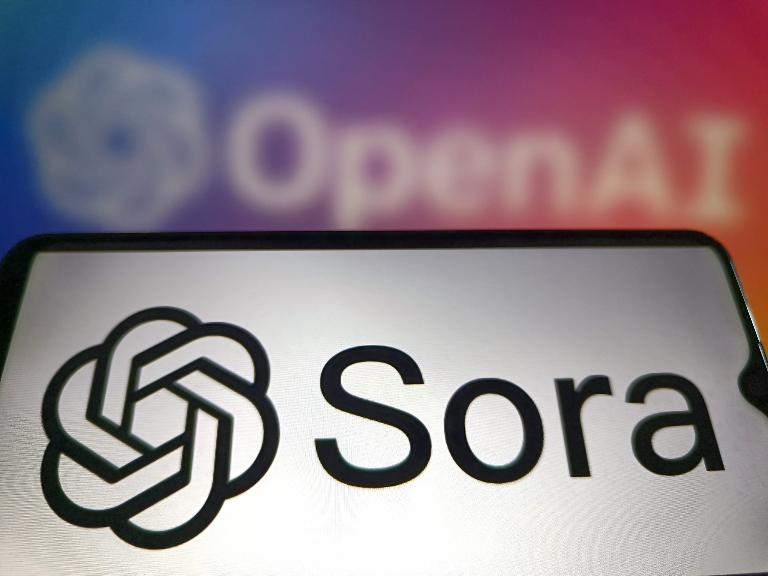 A smartphone screen displays the OpenAI "Sora" logo, with a blurred OpenAI logo in the colorful background, symbolizing AI innovation and branding.