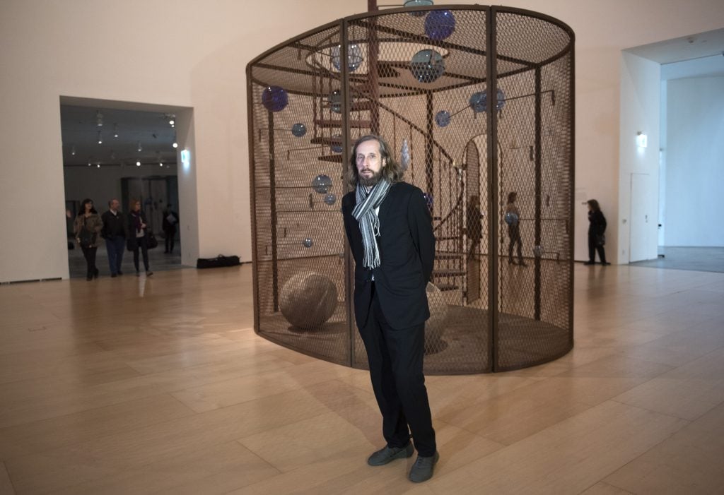 a man in a suit stands in front of a large sculpture in a cage structure in a art gallery