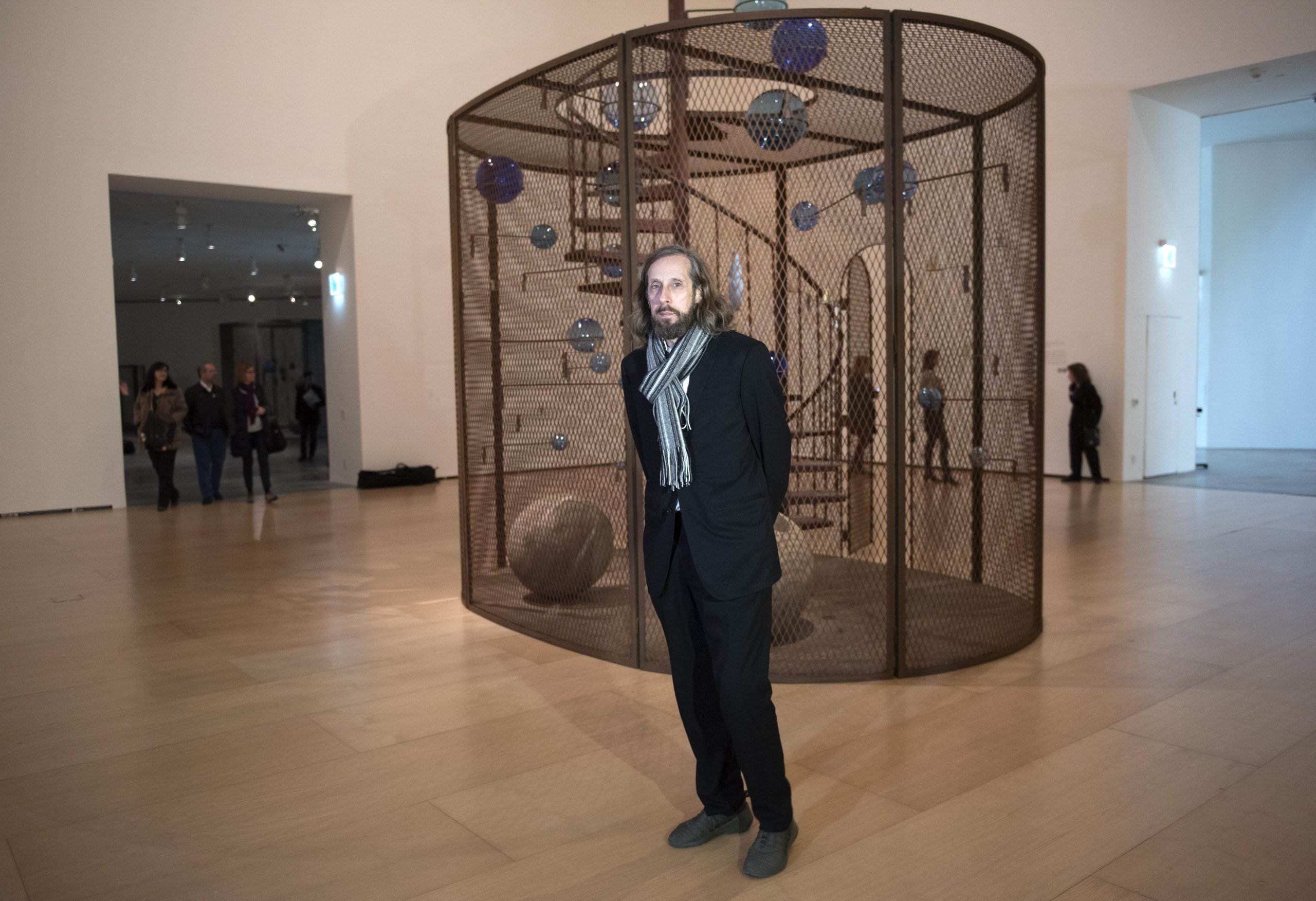 a man in a suit stands in front of a large sculpture in a cage structure in a art gallery