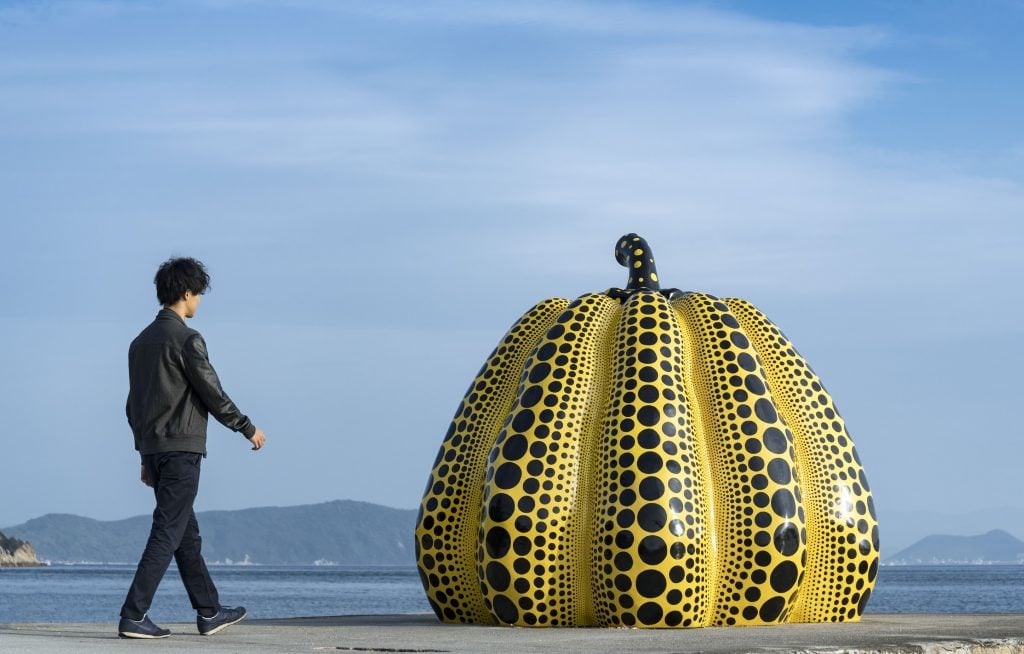 a figure is walking towards a yellow pumpkin sculpture with the sea behind them