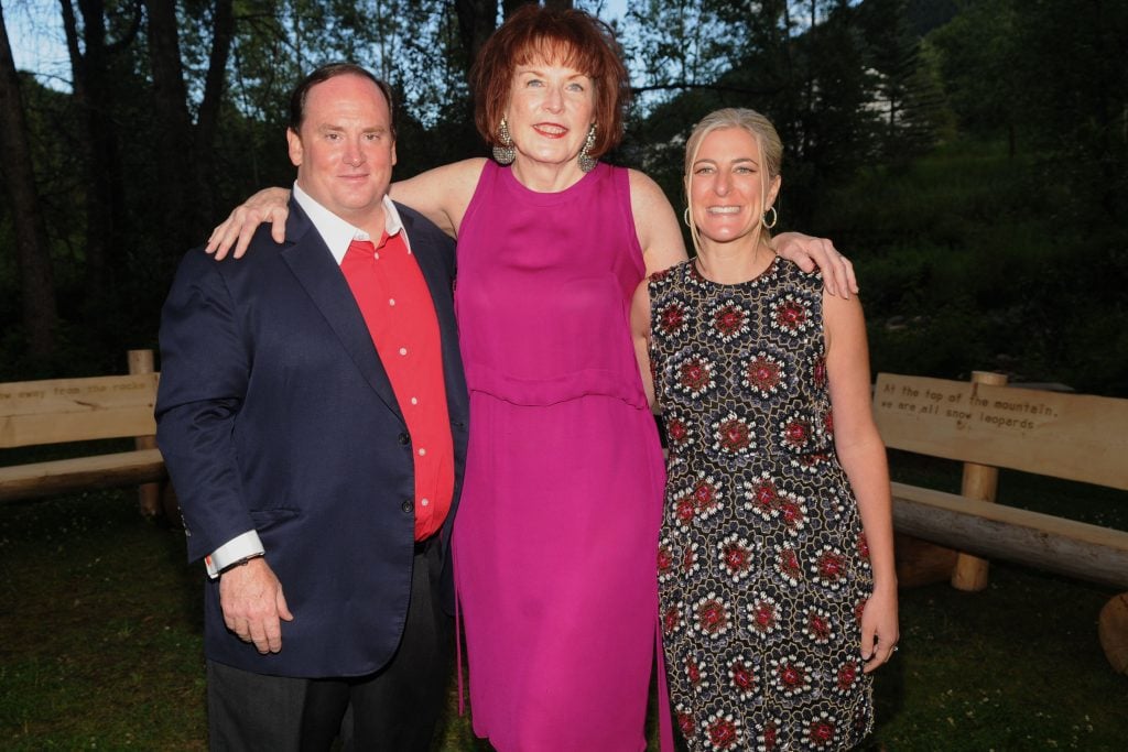 Three individuals pose outdoors, smiling warmly. A man in a red shirt, a tall woman in a pink dress, and another woman in a patterned dress.