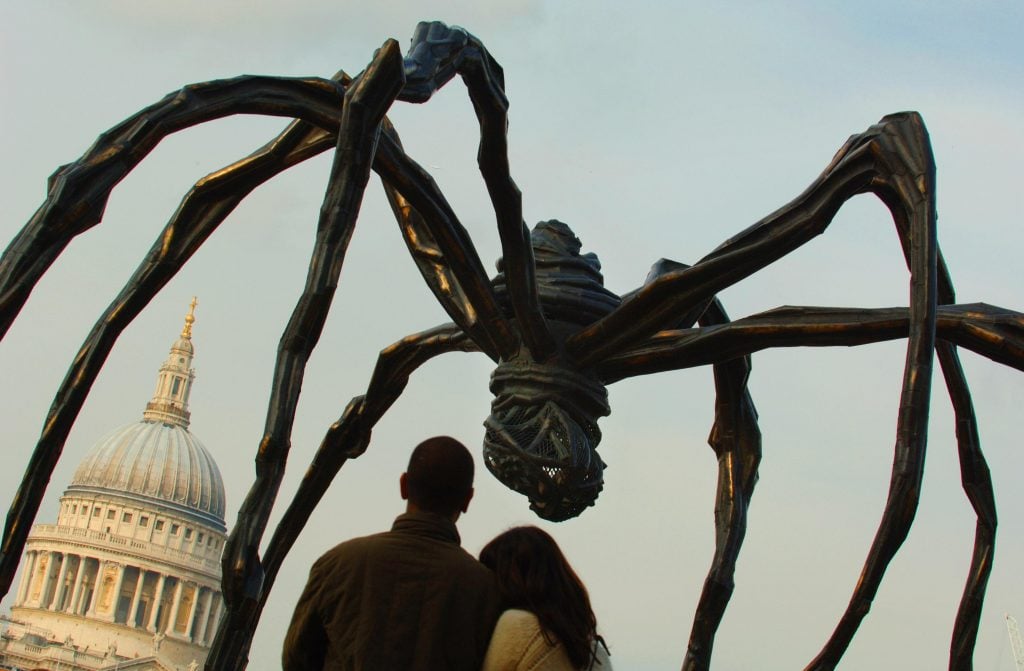 two people look up at a large spider sculpture outside with a large domed building in the background