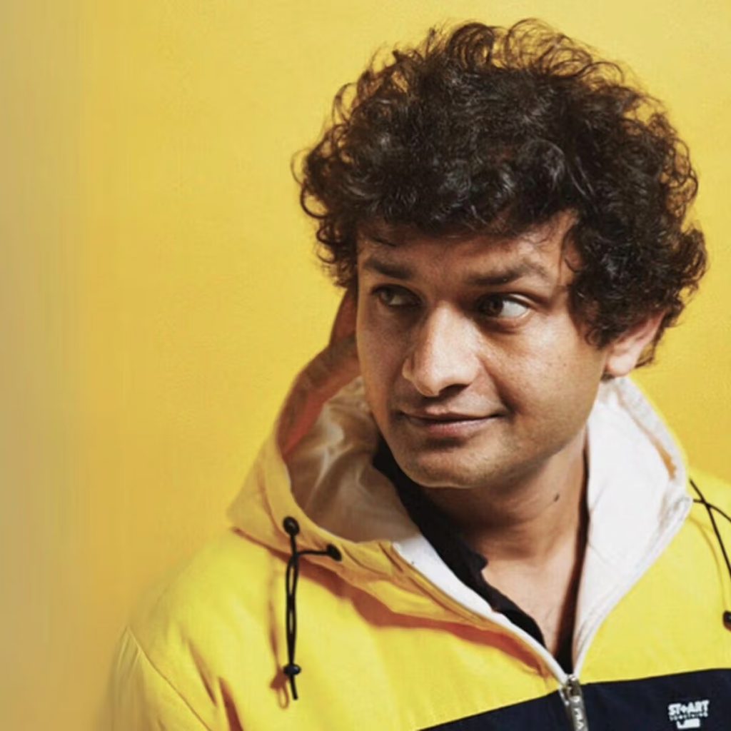 A photo of a young Indian man with thick curly hair wearing a hooded yellow jacket, standing in front of a yellow background. 