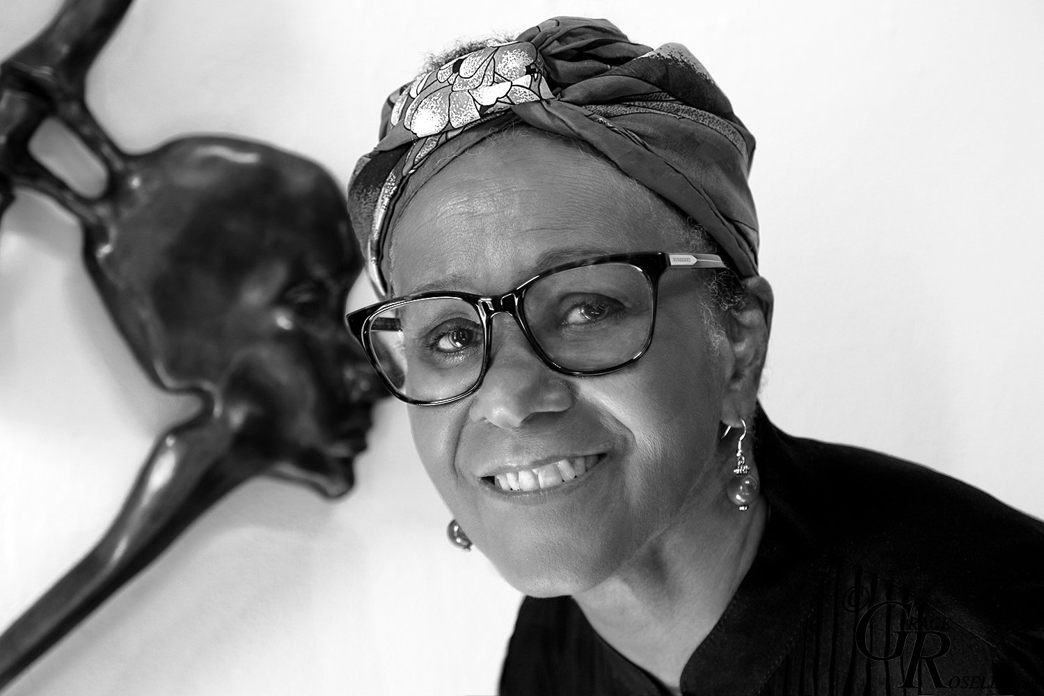 A black and white photograph of Helen Evans Ramsaran, an elderly Black woman with black-rimmed glasses and a turban. In the background, in soft focus, one of her bronze sculptures hangs on the wall.