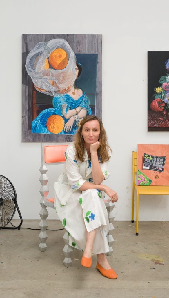 a white woman in a white dress with blue flowers sits on a chair in an artist studio with still life paintings behind her