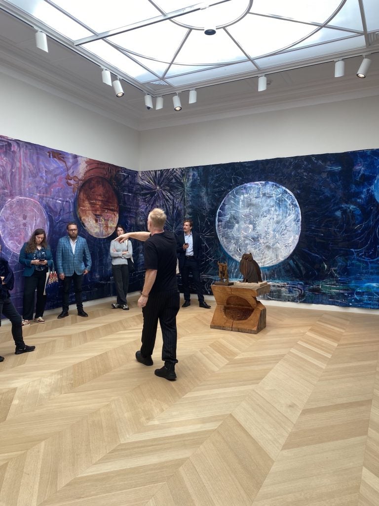 an image of the artist talking to visitors in front of a large room-size painted mural