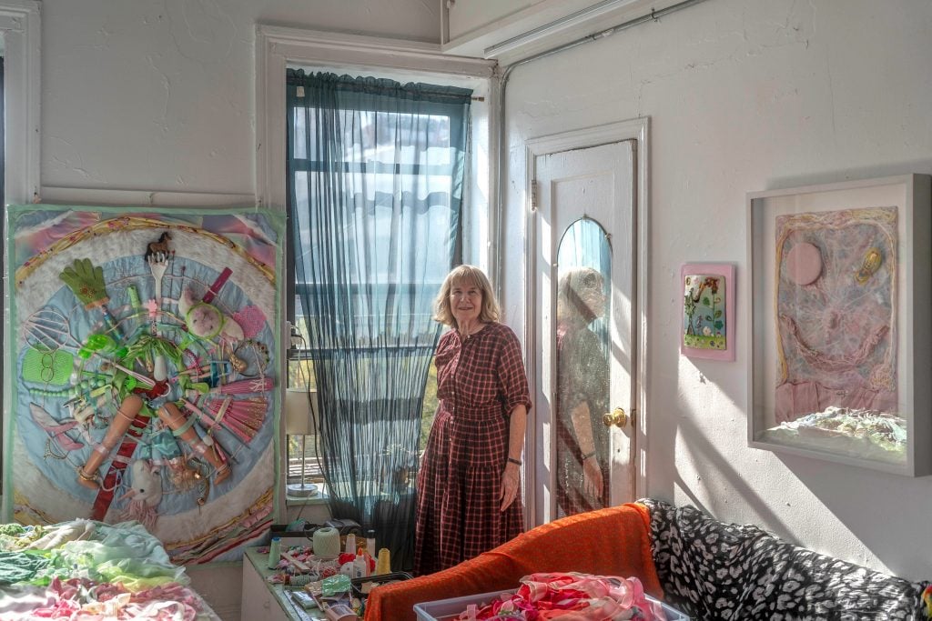 an older white woman with medium-length gray hair stands inside an apartment with art works visible behind her