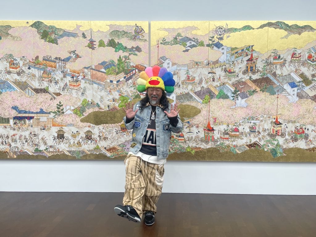 A chubby Asian man in glasses donning a colorful flower hat posed in front of a huge painting in gold and pink. The man is smiling and waving with both of his hands.