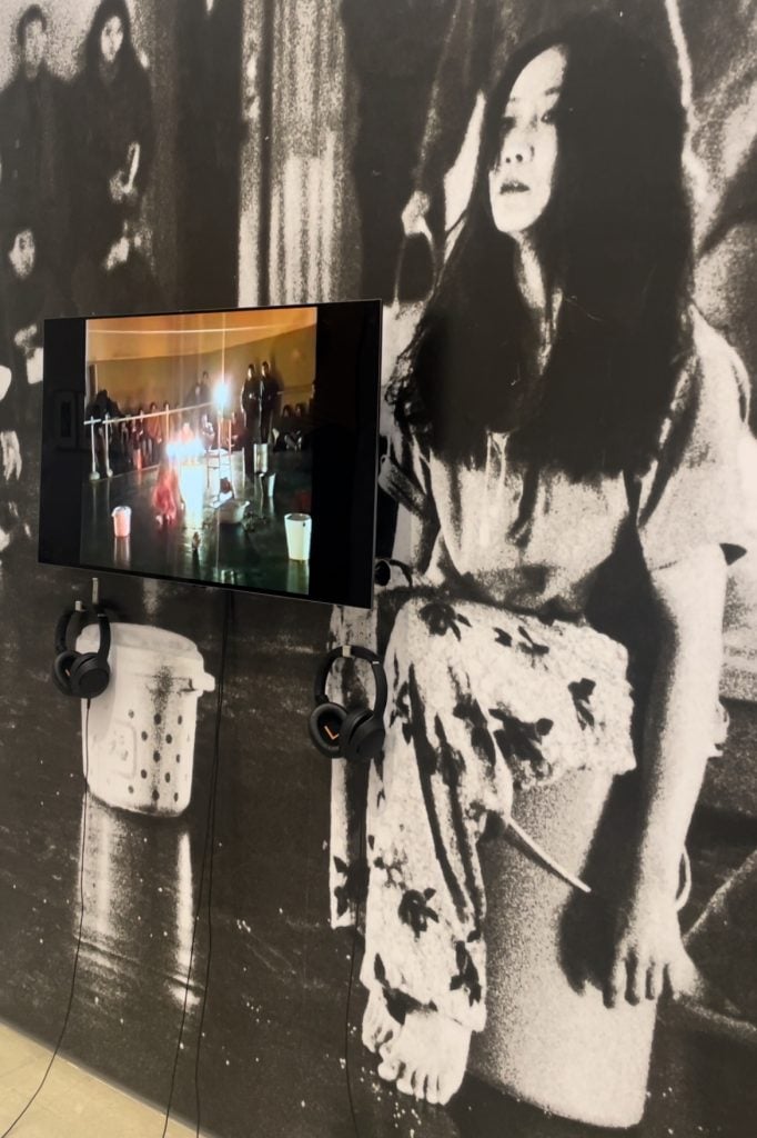 A video screen on the left, showing a performance that sees a woman with long black hair jumping around. The screen is attached to a wall covered by a large black and white photo of a woman in long dark hair, dressed in a white dress, sitting on a bucket.