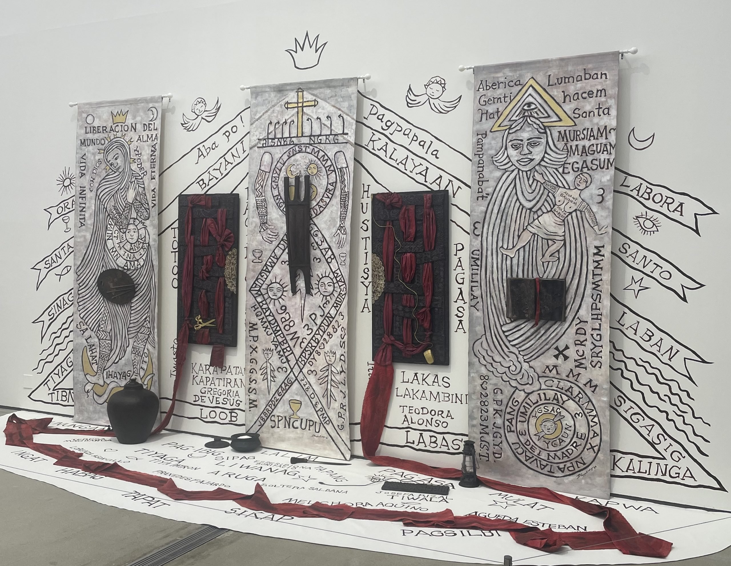 A mixed-media installation featuring four vertical banners with intricate hand-drawn designs and text in Filipino and Spanish, incorporating religious and cultural symbolism. Each banner is adorned with sculptures and three-dimensional elements such as red fabric and black objects. Surrounding the banners is a floor and wall installation with additional text and imagery, creating an immersive environment.