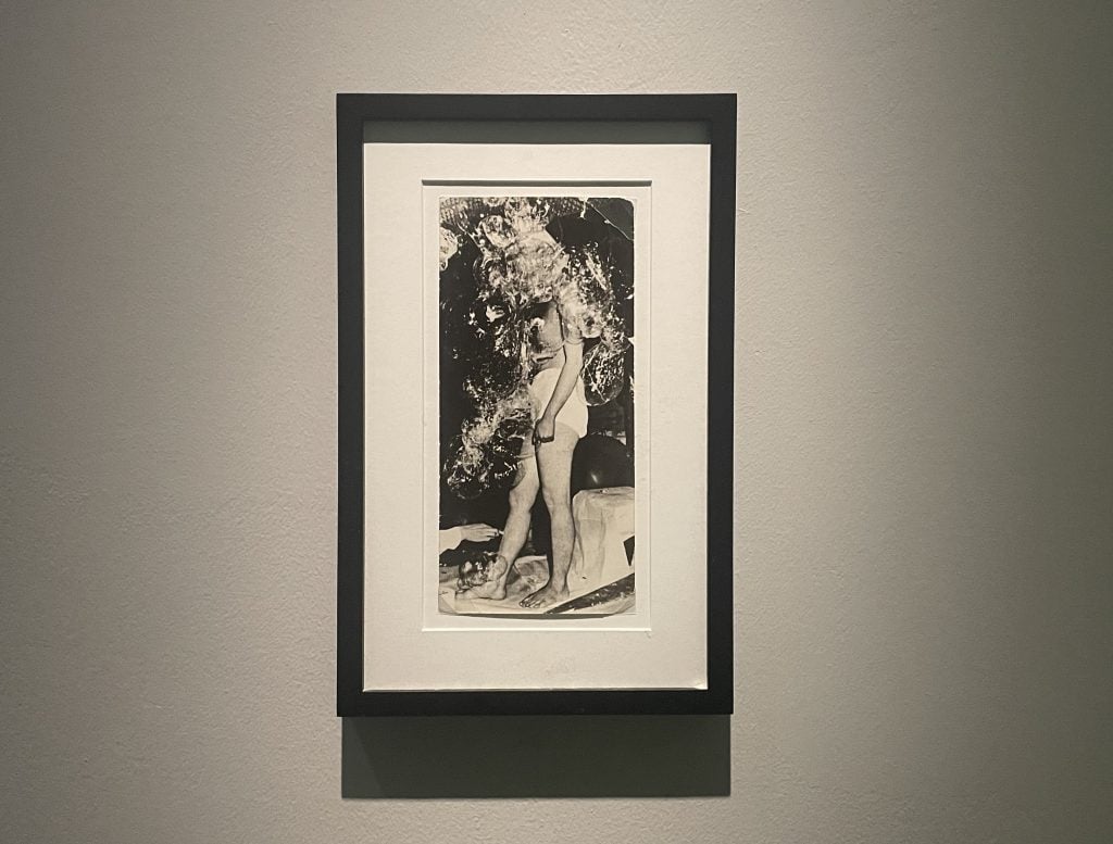 A framed black and white photograph of a woman with a naked upper body and white shorts, covering herself in transparent bubbles