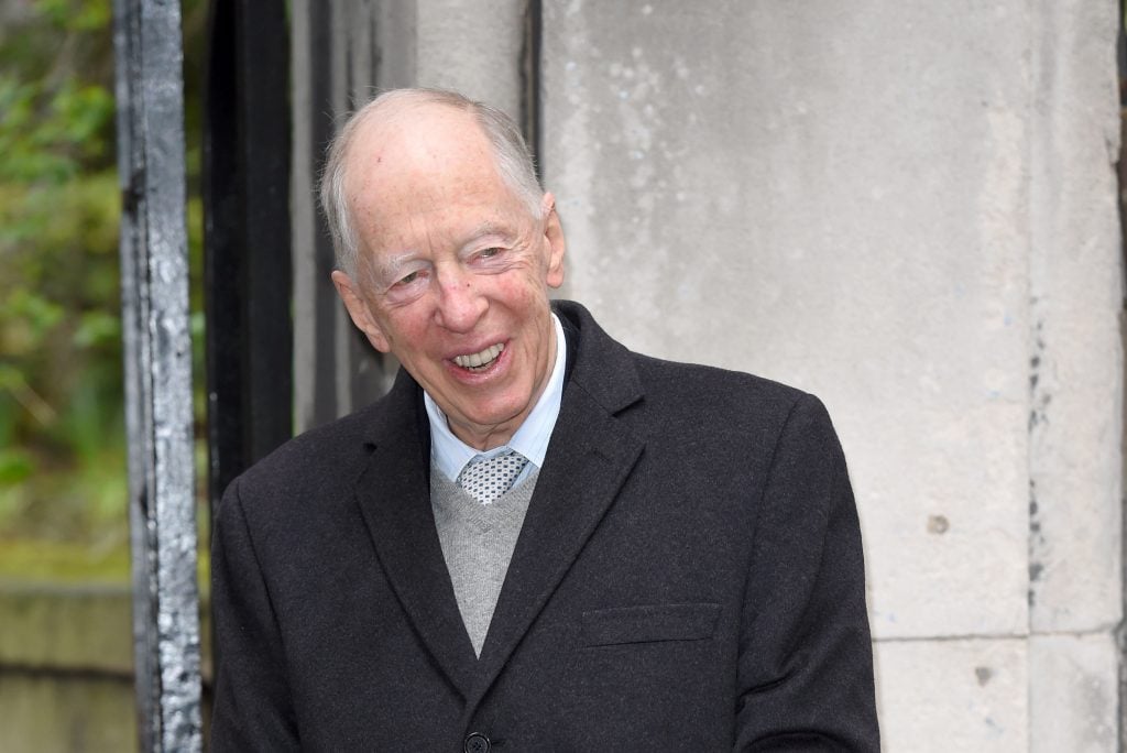 Jacob Rothschild. A balding, elderly white main in a suit smiling against the outside of a stone building. 