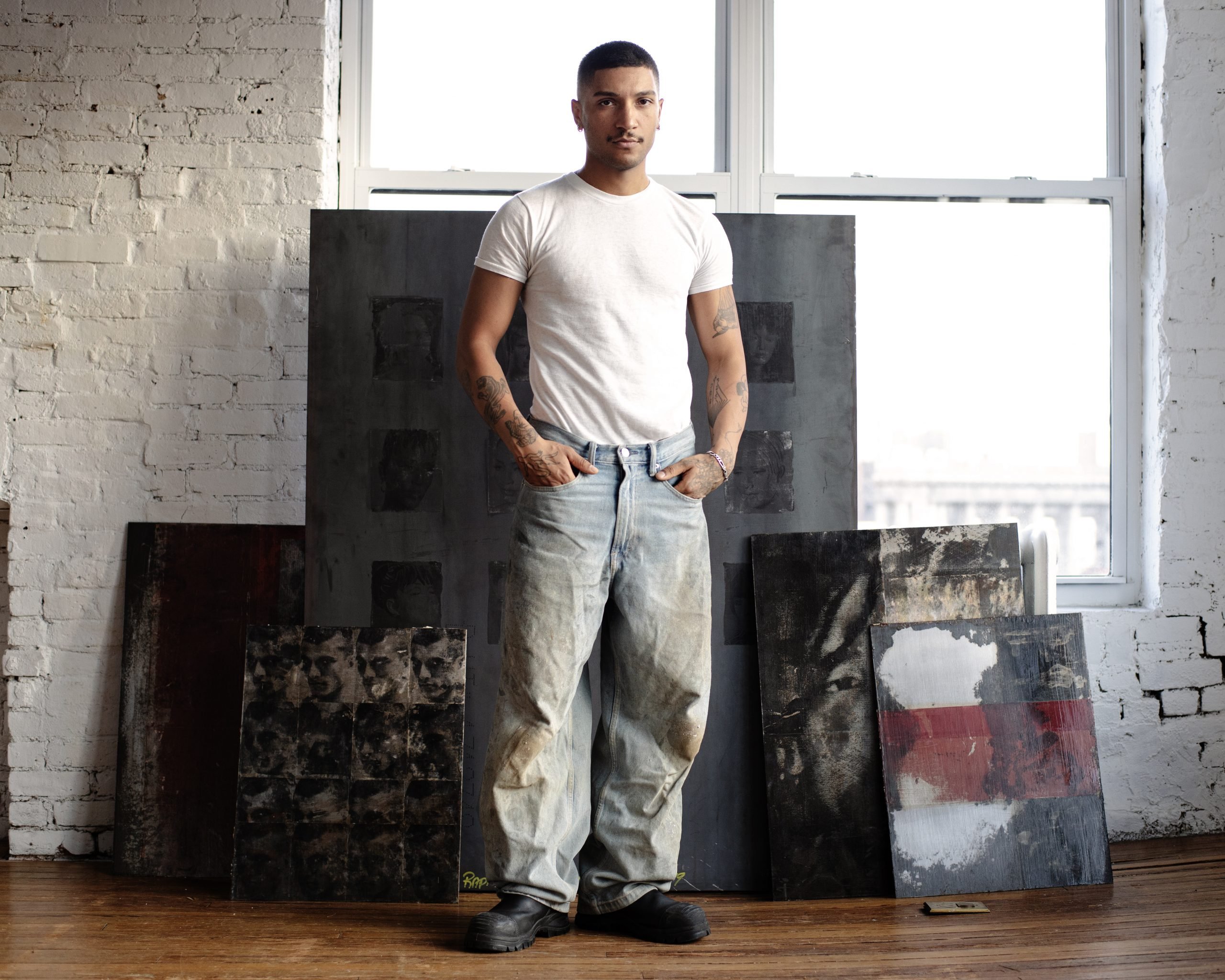 a young man with closecropepd black hair is wearing a white t shirt and jeans and is in a brick studio with the walls painted white in front of art