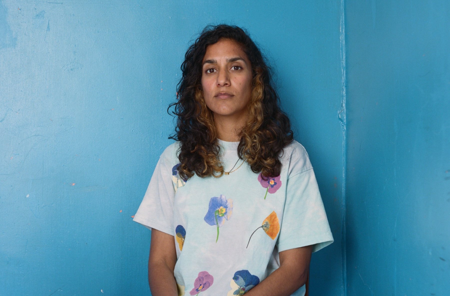 a woman with long dark curly hair in a white tee shirt sits against a bright blue wall