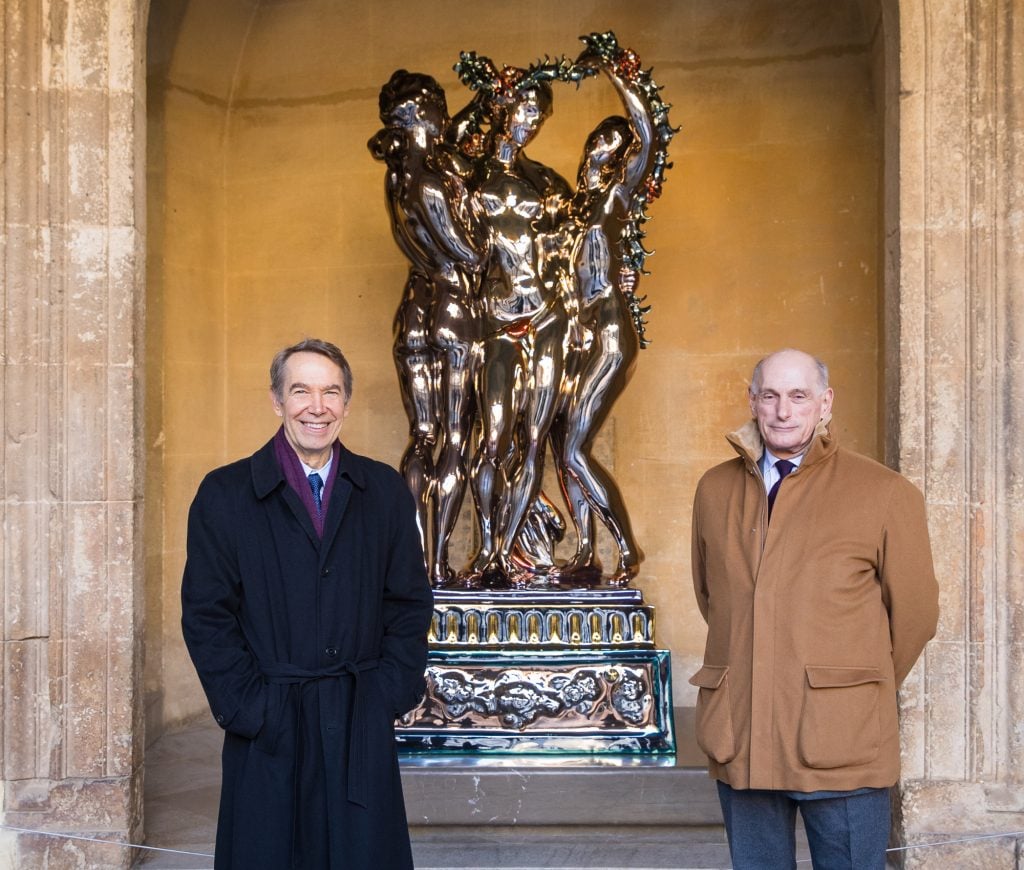 two men in coats stand beside a large very shiny sculpture of three female figures embracing each other and holding up a garland that is installed in a classical niche