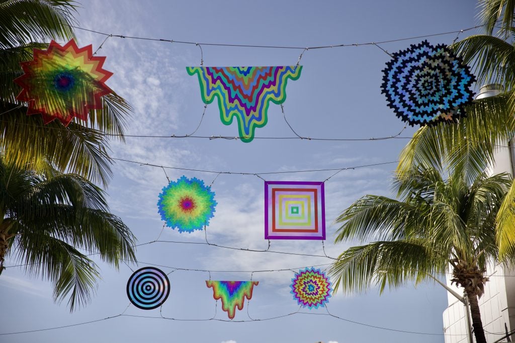 Jen Stark, Sundial Spectrum (2024). The seventh work in Elevate Española, a public art series on Española Way in Miami Beach. A series of translucent rainbow colored artworks hanging above the street, suspended between palm trees, are seen from below against a blue sky.