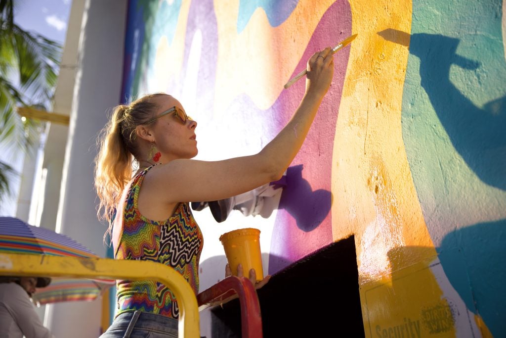 Jen Stark painting Sundial Spectrum (2024). She is a young blonde woman in sunglasses and a rainbow and black and white swirly striped body suit with a blonde ponytail, standing on a lift to paint a rainbow mural on a wall in the sunshine. 