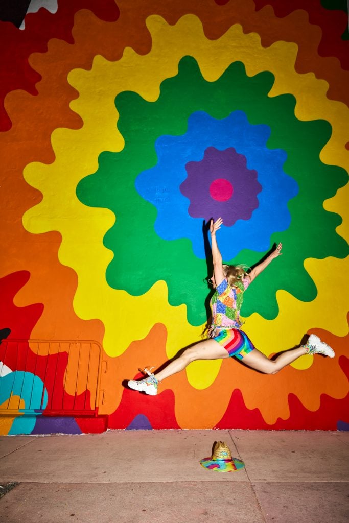 Jen Stark with her colorful mural Sundial Spectum (2024). She is a young woman in a rainbow colored short set, leaping in front of a matching mural of undulating concentric forms. 