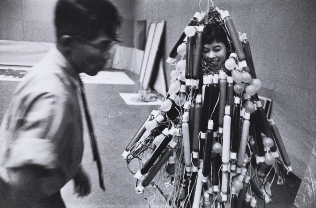 a black and white archival photograph of a woman wearing a "dress" that is a large mass of neon light emitters in different colours, we can't se the colours but we can assume the whole thing is very bright and colorful, she is swamped in the light and we can only just make out her face