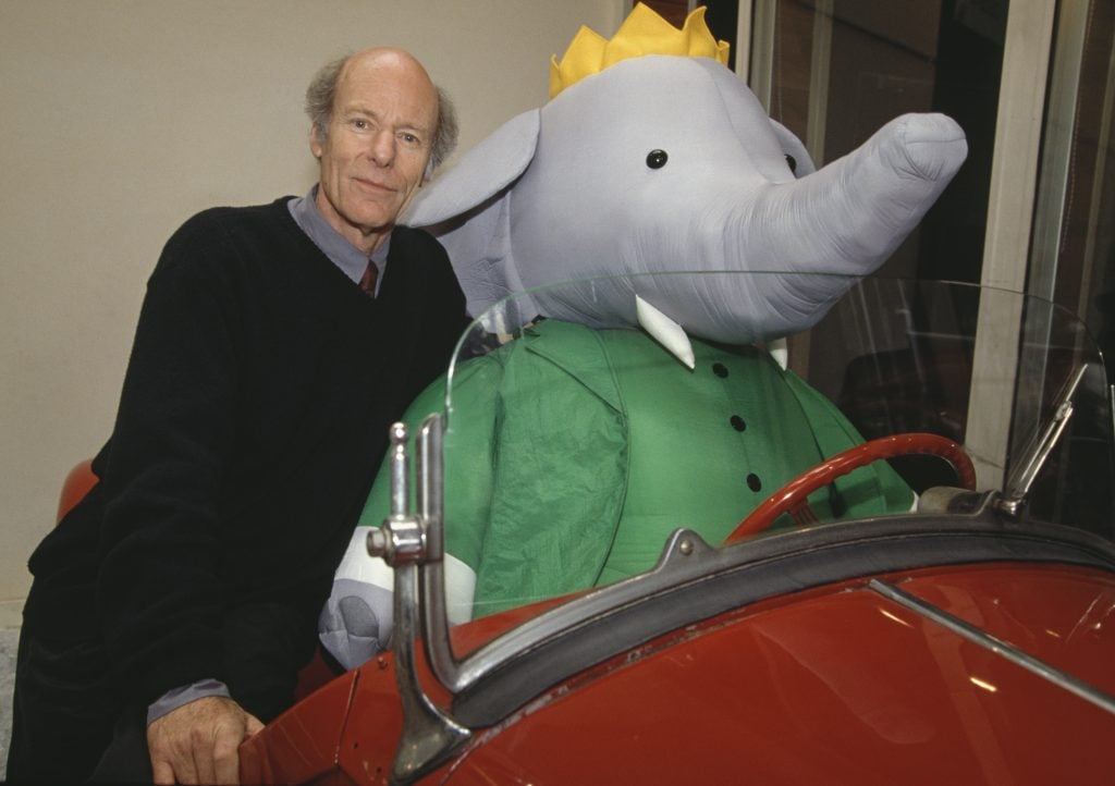 French cartoonist Laurent de Brunhoff in Paris in 1992 with a stuffed Babar the Elephant, celebrating the 60th anniversary of the famous character, created by his father, Jean de Brunhoff. He is an elderly white man sitting behind a giant stuffed elephant in a green coat and gold crown in an old-fashioned red convertible. 