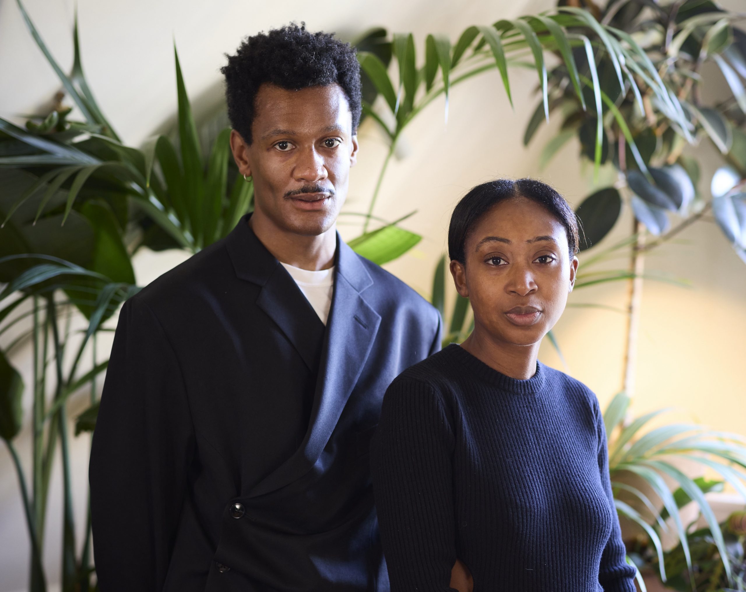 A man and woman posing in dark clothing in a lamp-lit room, in front of a plant