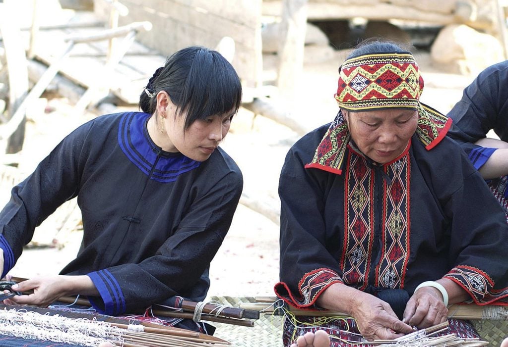 two women side-by-side do traditional weaving techniques, one peers over to look more closely at what the other is doing