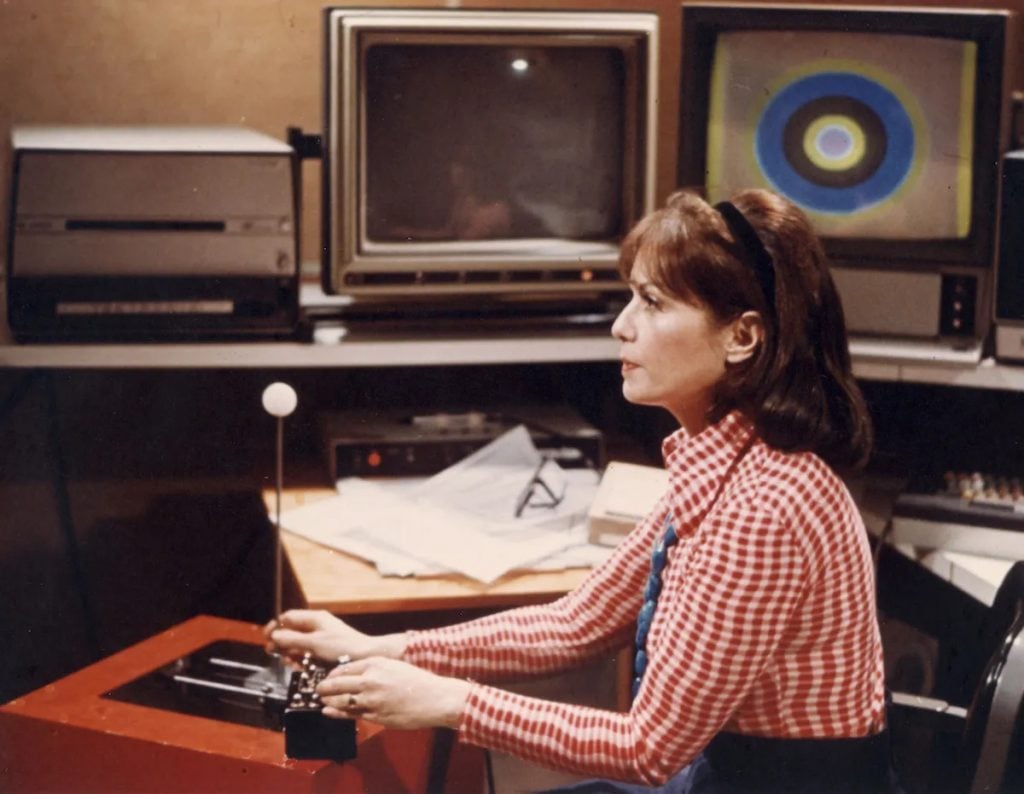 Lillian Schwartz (ca. 1975). A young woman in a red and white shirt with a printed pattern and dark hair pulled back by a black headband sits in profile at the controls at a computing device, with screens and other machines behind her. 