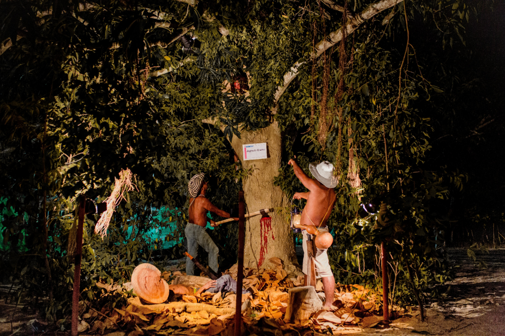 a brightly lit staged exterior scene of two men posed cutting down a tree that appears to be bleeding at the place it was cut into by an axe