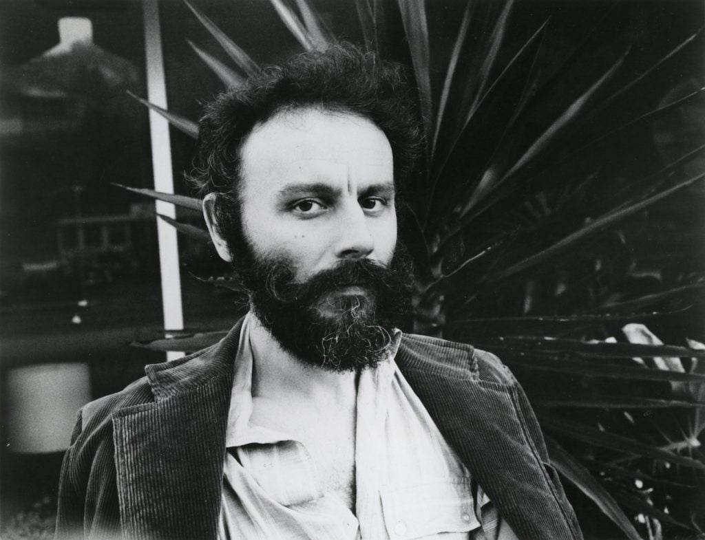 A black and white photo of a young Lucas Samaras, a young artist with curly dark hair and a bushy beard, wearing a rumpled white collared shirt and a corduroy blazer. He is standing in front of a large palm leaf. 