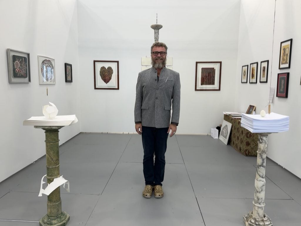 Marco Di Giovanni with his work at Untitled Art in Miami Beach, shown by Il Chiostro Arte & Archivi, Saronno, Italy. He is a middle aged man with a beard in a gray shirt and black pants standing in a gallery booth with white walls hung with art, and two column sculptures to either side of him. 