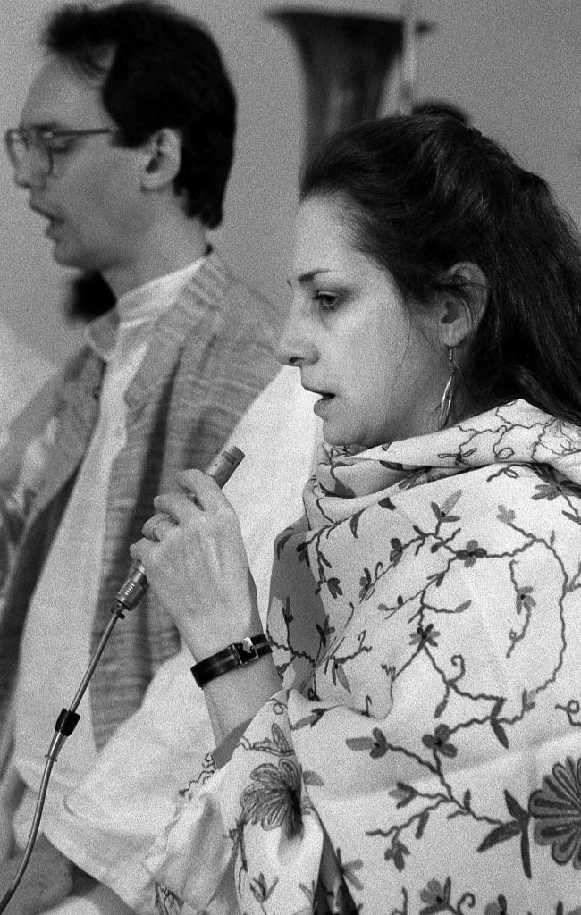 American light artist and designer Marian Zazeela (ca. 1991). It's a black and white photo of the artist in profile, speaking into a small microphone. She is wearing an Indian looking robe with a floral embroidery on it.