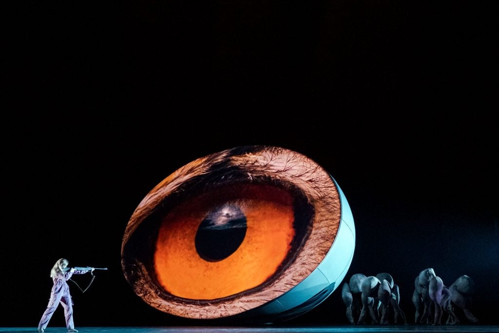 a dancer stands on a stage in a one piece in front of a large projection of an owl's eye