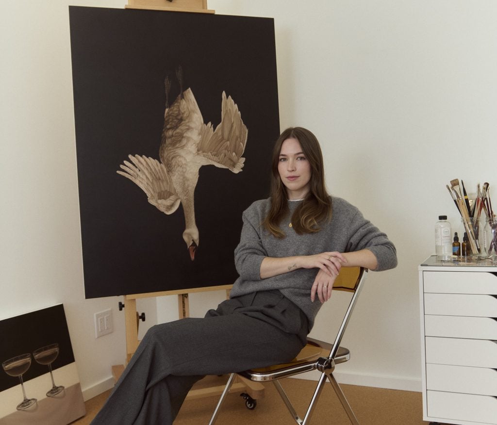 artist Meghann Stephenson, a young white woman with medium length brown hair, seated in a chair wearing a gray sweater and trousers, next to a painting of a swan against a dark black background