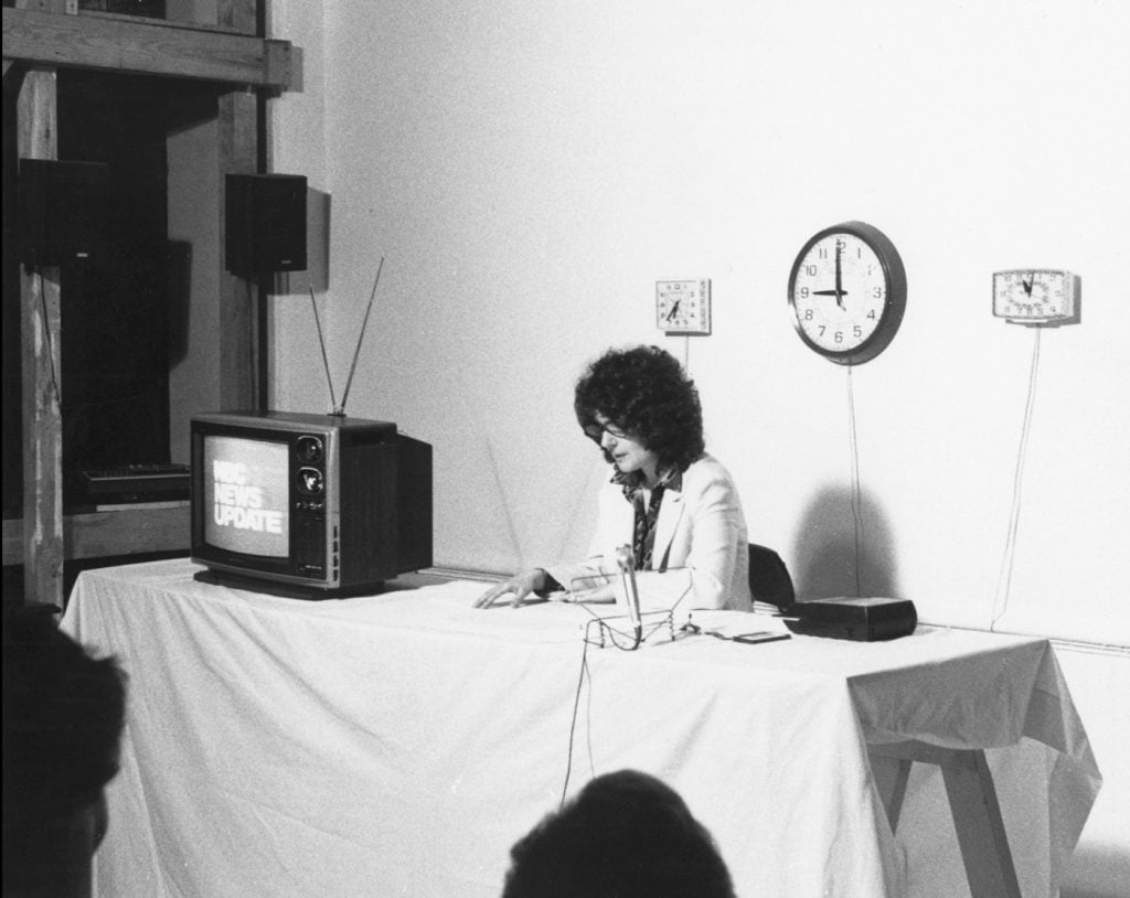 Mimi Smith, A Reading of Two Drawings 1978 Franklin Furnace. A black and white photo of the artist sitting at a table in front of a round clock next to a vintage television with an antenna, reading the transcript of the nightly news aloud.