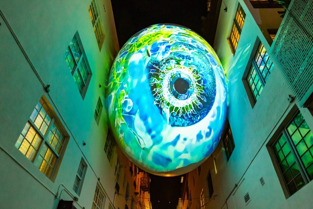 [dNASAb], Faux Ecologies + Augmented Visions of the Micro-verse, on view in “No Vacancy, Miami Beach” on "The Orb" at the Betsy, Miami Beach. A projected blue and green abstract artwork is seen on a spherical egg-shaped skybridge connecting two buildings.