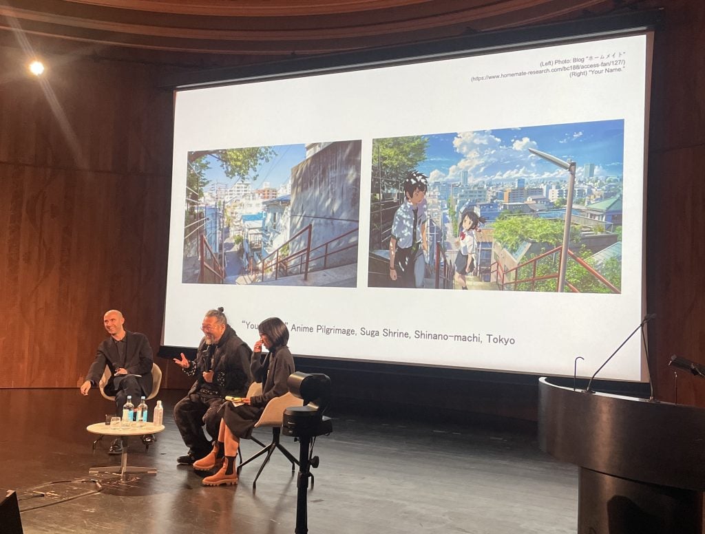 Three people sitting on stage in front of a large screen, showing images of a still shot of an anime and the other a photo of an actual location.