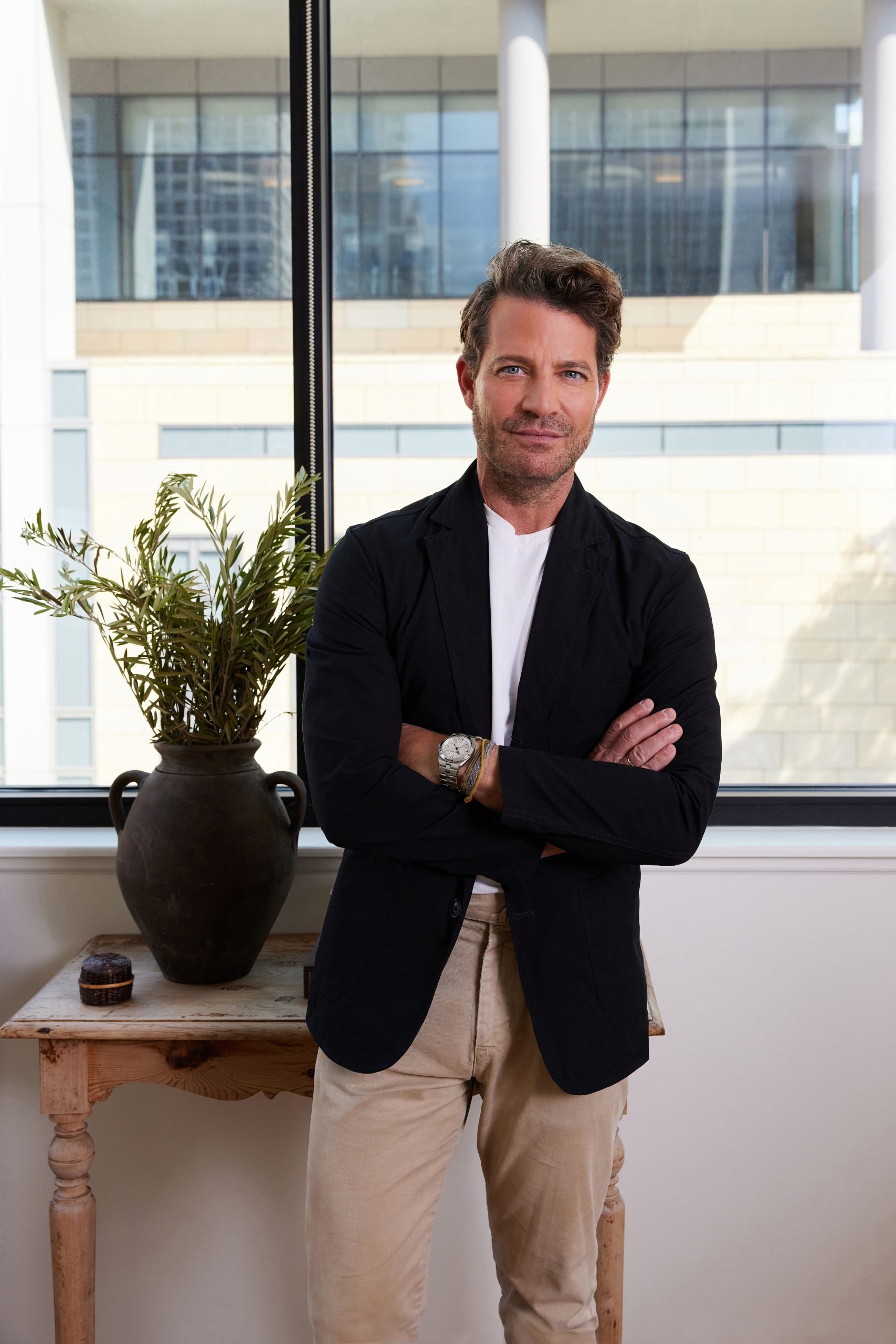 a photograph of a white man with brown hair wearing tan pants and a black blazer. The man pictured in the decorator Nate Berkus