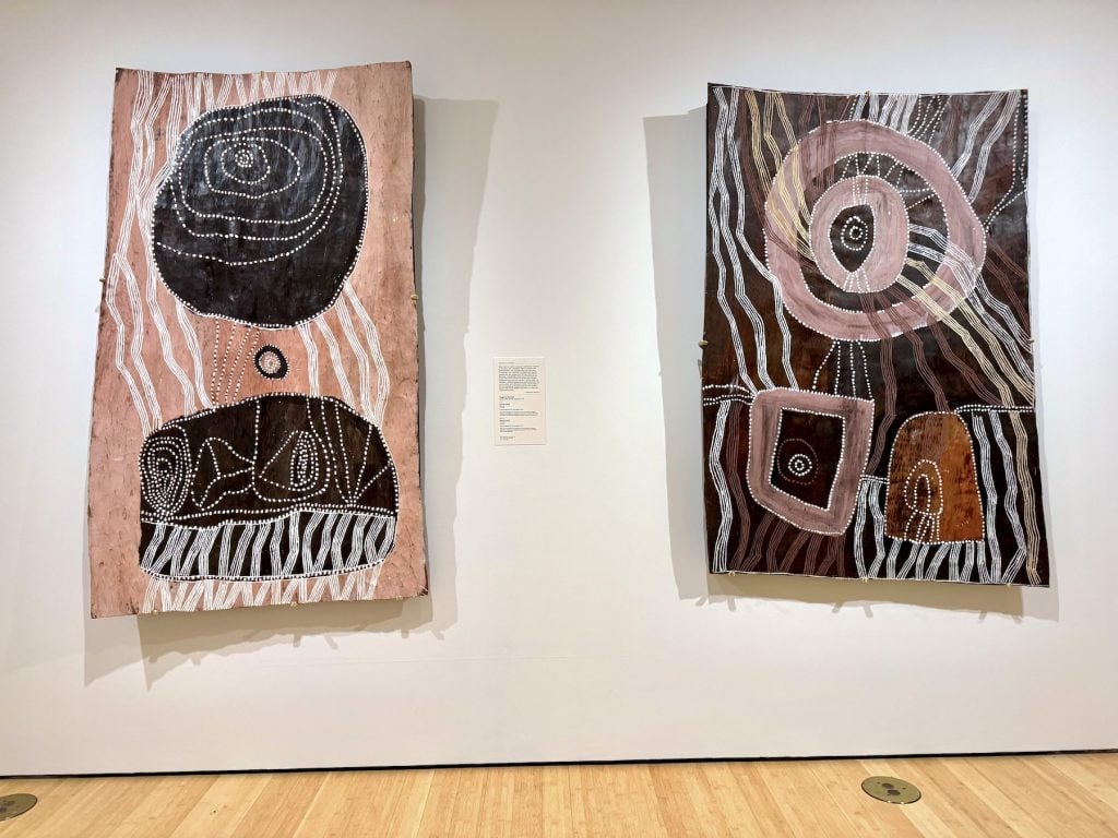 Two pieces of Indigenous Australian bark art are displayed on a white gallery wall