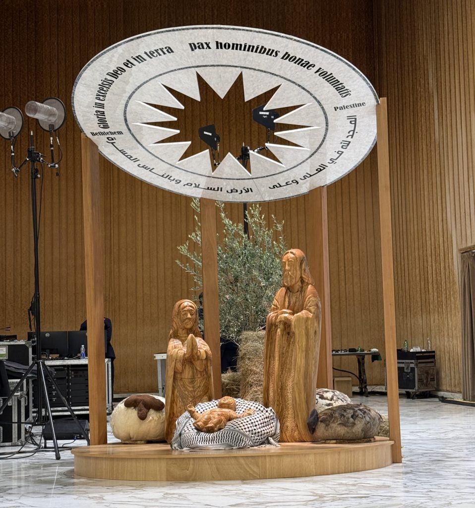 a wooden nativity shows mary and jesus standing in prayer on either side of a wooden baby resting on a Palestinian keffiyeh, overhead it is sheltered by a white disc with writing written around the outer rim and a star cut into the middle