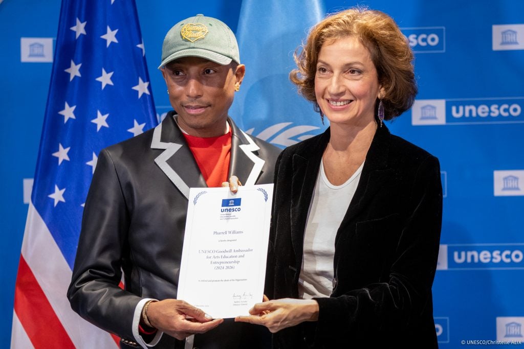 Pharrell Williams and Audrey Azoulay hold a piece of paper and smile