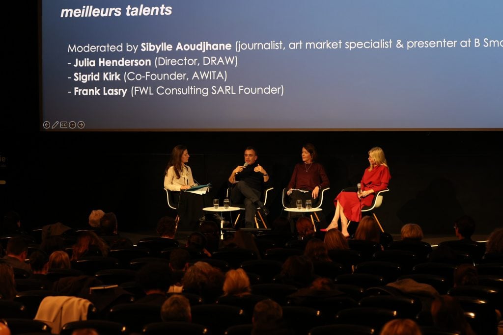 4 people sit in chairs on stage with a large projection screen behind them