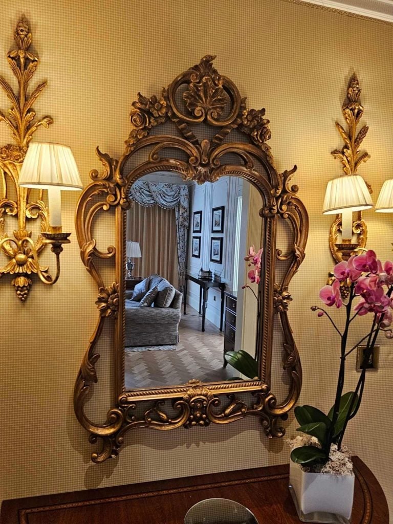 photograph of a large mirror encased in a hand carved ornamental frame with two lampshades on either side above a little wooden table with a large red flower in a vase